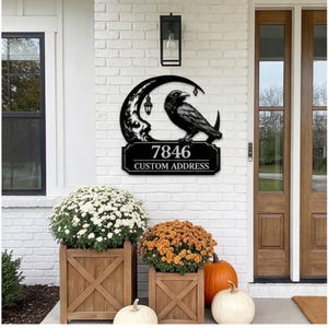 May include: Black metal address sign featuring a raven perched on a crescent moon with lanterns. The sign displays the number 7846 and the words "CUSTOM ADDRESS". The sign is mounted on a white brick wall near a wooden door.