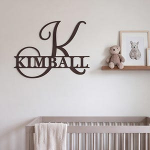 Könnte beinhalten: Ein dunkelbraunes Wand-Schild mit einem großen, stilisierten Großbuchstaben K, wobei der Name KIMBALL in den horizontalen Querbalken integriert ist. Das Schild ist mit einer Schreibschrift für den Anfangsbuchstaben und einer serifenlosen Schrift gestaltet.