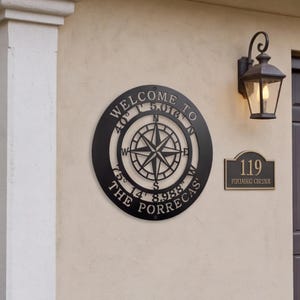 Pode incluir: Uma placa de parede circular em metal preto com um design central de rosa dos ventos. O texto diz WELCOME TO, THE PORRECAS, e as coordenadas 40° 1' 5.016" N 75° 14' 8.988" W. A placa está montada numa parede clara junto a uma placa numérica.