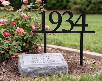 May include: A black metal garden stake featuring the numbers 1934 in a bold serif font, positioned behind a rectangular grey granite memorial stone with engraved text, set in a garden bed with pink rose bushes and green grass.