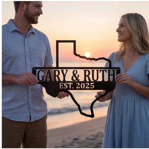 May include: Black metal Texas-shaped sign with the names "GARY & RUTH" and the year "EST. 2025" cut out. The sign is held by two people on a beach at sunset. The sign is a personalized home decor item.