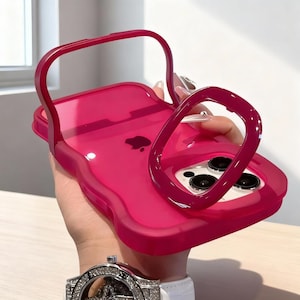 May include: A bright pink phone case with a handle and a ring grip. The case has a transparent design, showcasing the phone's camera lenses and the Apple logo. The handle and ring grip are the same shade of pink.