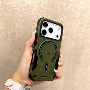 May include: An olive green phone case with black detailing. The case has a built-in stand and a circular design with the text "TECH & DESIGN". It features a charging symbol and is designed to protect the phone.