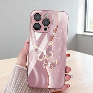 May include: A pink phone case with a butterfly design. The case has a glossy finish and features two butterflies with gold accents. The case has a camera cutout and a curved design.
