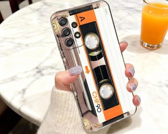 Puede incluir: Una funda transparente para teléfono con un diseño de cinta de cassette retro. La funda tiene una combinación de colores naranja, negro y blanco con el texto "CHF90" y "A" visibles. La funda se sostiene en una mano.