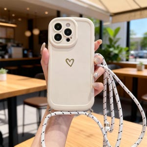 May include: A cream-coloured phone case featuring a gold heart design and a matching lanyard. The case has a raised border around the camera and a white and black patterned strap. The phone case is held in front of a blurred background.
