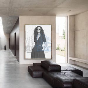 Może przedstawiać: Oprawione dzieło sztuki przedstawiające kobietę w okularach przeciwsłonecznych, z niebieskim nadrukiem architektonicznym. Grafika wisi w nowoczesnym, minimalistycznym wnętrzu z ciemnobrązową skórzaną sofą modułową. Pomieszczenie ma betonowe ściany i podłogi.