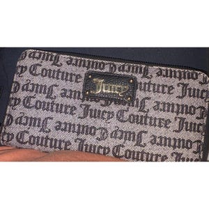 May include: A rectangular wallet featuring a repeating pattern of the words "Juicy Couture" in black script on a light brown background. The wallet has a black leather patch with the word "Juicy" in gold lettering.