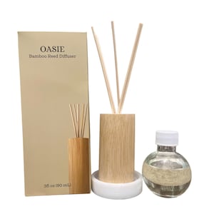 May include: OASIE Bamboo Reed Diffuser set including a tall beige rectangular box with text, a cylindrical light wood diffuser holder with three reeds, a white circular base, and a clear glass bottle with a white cap containing 3 fl oz of liquid.