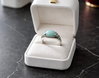 May include: A silver ring with a large, oval, light blue gemstone is displayed in a white jewelry box. The ring has green leaf accents. The box is open, revealing a white interior and a small gold crown on the lid.