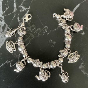 Puede incluir: Pulsera de abalorios plateada con abalorios de tetera y taza de té. La pulsera tiene una mezcla de cuentas redondas y texturizadas. Los abalorios y las cuentas están ensartados con una cadena y un cierre plateados.