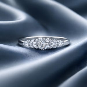 Könnte beinhalten: Ein silberner Ring mit sechs runden, klaren Edelsteinen. Der Ring liegt auf einem blauen Satin-Stoff, wodurch ein Kontrast zwischen den funkelnden Steinen und dem weichen Hintergrund entsteht. Die Edelsteine sind in einer Reihe angeordnet, mit einem größeren Stein in der Mitte.