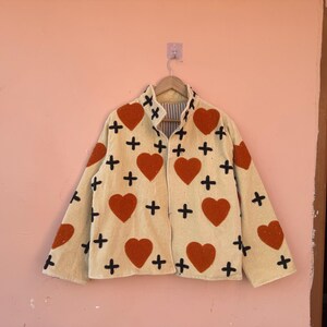 May include: Cream-colored jacket with large red heart and black cross patterns. The jacket is hanging on a wooden hanger against a pink wall. The jacket has a striped lining.