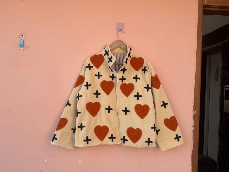 May include: Cream-colored jacket with large red heart and black cross patterns. The jacket is hanging on a wooden hanger against a pink wall. The jacket has a striped lining.