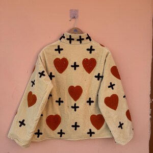 May include: Cream-colored jacket with red heart and black cross appliqu&eacute;s. The jacket has a high collar and long sleeves. The hearts are evenly spaced across the front and sleeves. The jacket is hanging on a metal hanger.