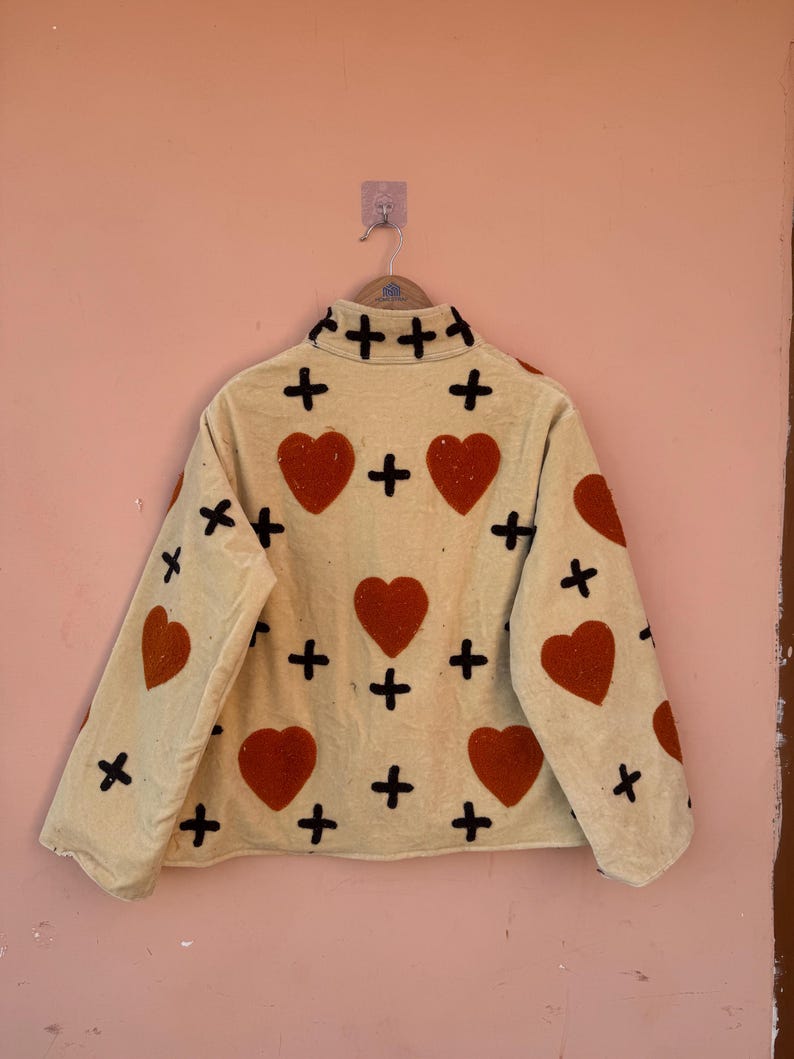 May include: Cream-colored jacket with red heart and black cross appliqu&eacute;s. The jacket has a high collar and long sleeves. The hearts are evenly spaced across the front and sleeves. The jacket is hanging on a metal hanger.