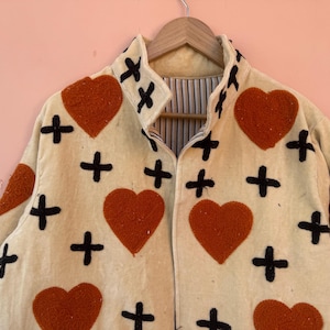 May include: Cream-colored jacket with a repeating pattern of large orange hearts and black crosses. The jacket is hanging on a wooden hanger against a pink background. The jacket has a collared neckline.