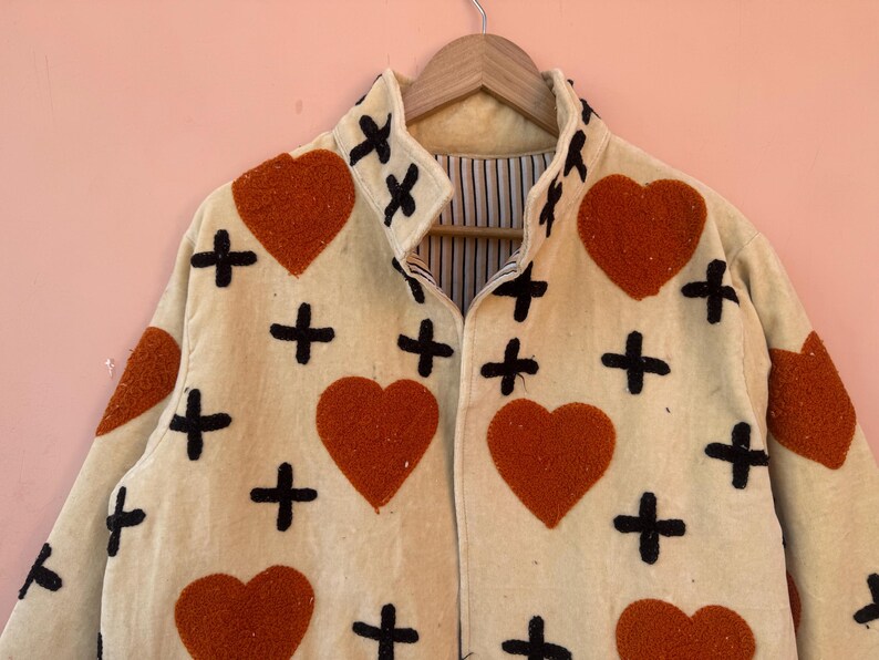 May include: Cream-colored jacket with a repeating pattern of large orange hearts and black crosses. The jacket is hanging on a wooden hanger against a pink background. The jacket has a collared neckline.