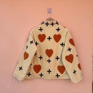 May include: Beige jacket with a high collar, adorned with large red hearts and black cross patterns. The jacket is hanging on a pink wall. The jacket's design is repeated on the sleeves and back.