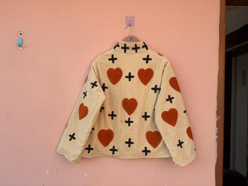 May include: Beige jacket with a high collar, adorned with large red hearts and black cross patterns. The jacket is hanging on a pink wall. The jacket's design is repeated on the sleeves and back.