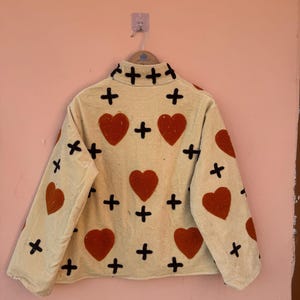 May include: A cream-colored jacket with a high collar, adorned with large red hearts and black cross accents. The jacket is hanging on a hanger against a peach-colored wall. The jacket's design is repeated on the sleeves.