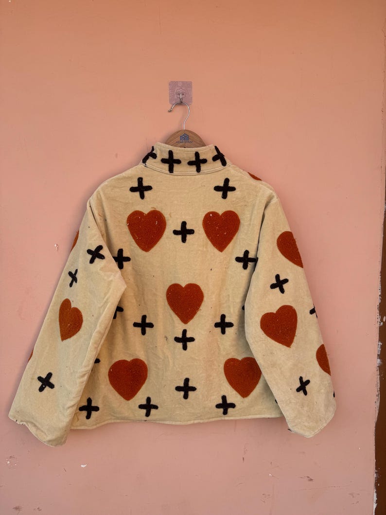 May include: A cream-colored jacket with a high collar, adorned with large red hearts and black cross accents. The jacket is hanging on a hanger against a peach-colored wall. The jacket's design is repeated on the sleeves.