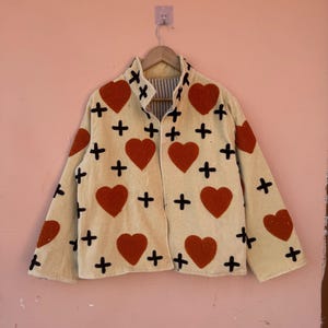 May include: Cream-colored jacket with red heart and black cross appliqu&eacute;s. The jacket is hanging on a wooden hanger against a pink wall. The jacket has a collared neckline and a striped lining.