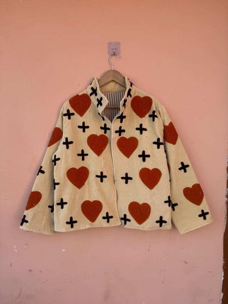 May include: Cream-colored jacket with red heart and black cross appliqu&eacute;s. The jacket is hanging on a wooden hanger against a pink wall. The jacket has a collared neckline and a striped lining.