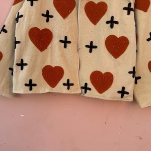 May include: Cream-colored jacket with large, rust-colored hearts and black cross accents. The jacket has long sleeves and a simple, open front design. The fabric appears to be a textured material.