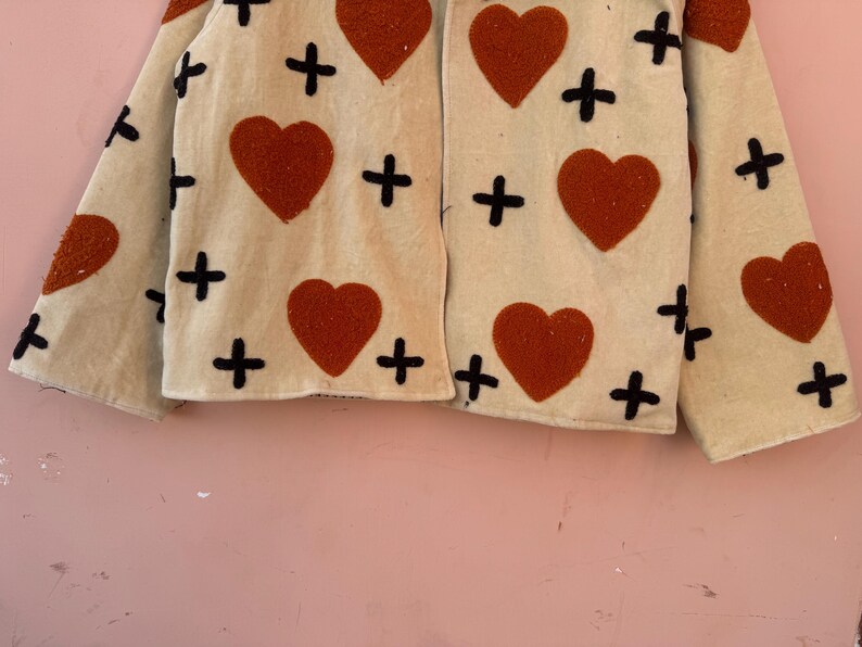 May include: Cream-colored jacket with large, rust-colored hearts and black cross accents. The jacket has long sleeves and a simple, open front design. The fabric appears to be a textured material.
