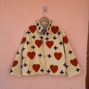 May include: Cream-colored jacket with large red heart appliqu&eacute;s and black cross accents. The jacket is hanging on a wooden hanger against a pink wall. The jacket has a collared neckline and long sleeves.