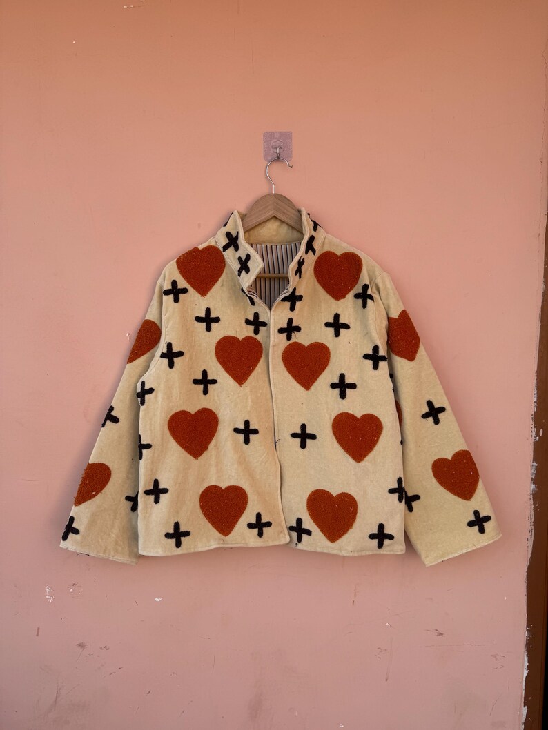 May include: Cream-colored jacket with large red heart appliqu&eacute;s and black cross accents. The jacket is hanging on a wooden hanger against a pink wall. The jacket has a collared neckline and long sleeves.