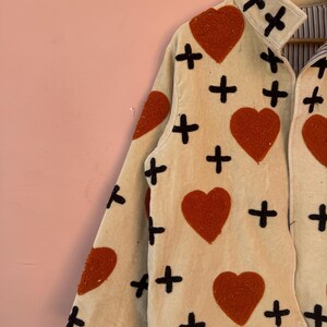 May include: Cream-colored jacket with large, textured, orange hearts and black cross symbols. The jacket has a collared neckline and long sleeves. The interior lining is striped.