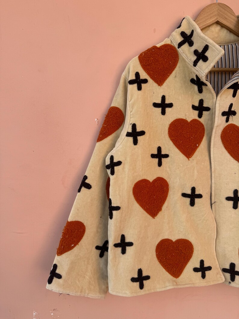 May include: Cream-colored jacket with large, textured, orange hearts and black cross symbols. The jacket has a collared neckline and long sleeves. The interior lining is striped.