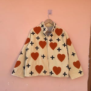 May include: Cream-colored jacket with a pattern of large red hearts and black crosses. The jacket is hanging on a wooden hanger against a pink wall. The jacket has a collared neckline and long sleeves.