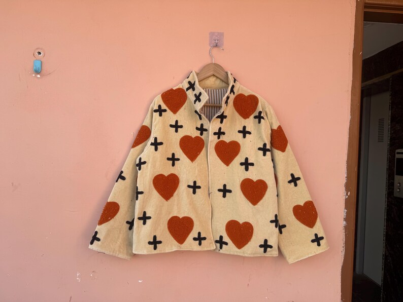 May include: Cream-colored jacket with a pattern of large red hearts and black crosses. The jacket is hanging on a wooden hanger against a pink wall. The jacket has a collared neckline and long sleeves.