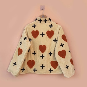 May include: Cream-colored jacket with a repeating pattern of red hearts and black crosses. The jacket is hanging on a wooden hanger against a light pink wall. The jacket has long sleeves and a collared neckline.