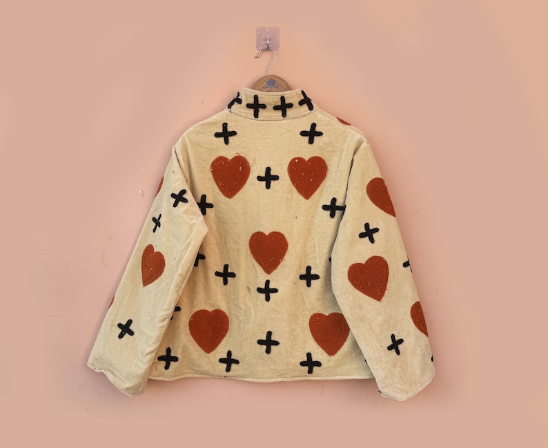 May include: Cream-colored jacket with a repeating pattern of red hearts and black crosses. The jacket is hanging on a wooden hanger against a light pink wall. The jacket has long sleeves and a collared neckline.