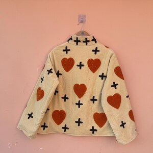 May include: A beige jacket with a heart and cross pattern. The jacket features large orange hearts and black crosses. The jacket is hanging on a pink wall.