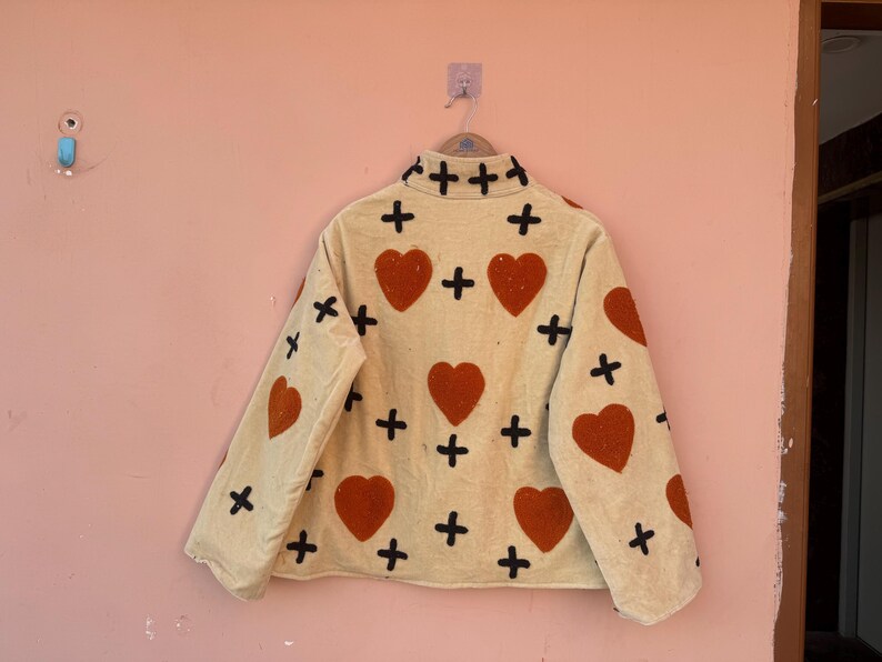 May include: A beige jacket with a heart and cross pattern. The jacket features large orange hearts and black crosses. The jacket is hanging on a pink wall.