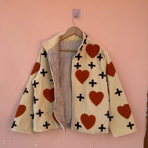 May include: Cream-colored jacket with red heart and black cross appliqu&eacute;s. The jacket has a collared neckline and a striped interior lining. It is hanging on a wooden hanger against a pink wall.