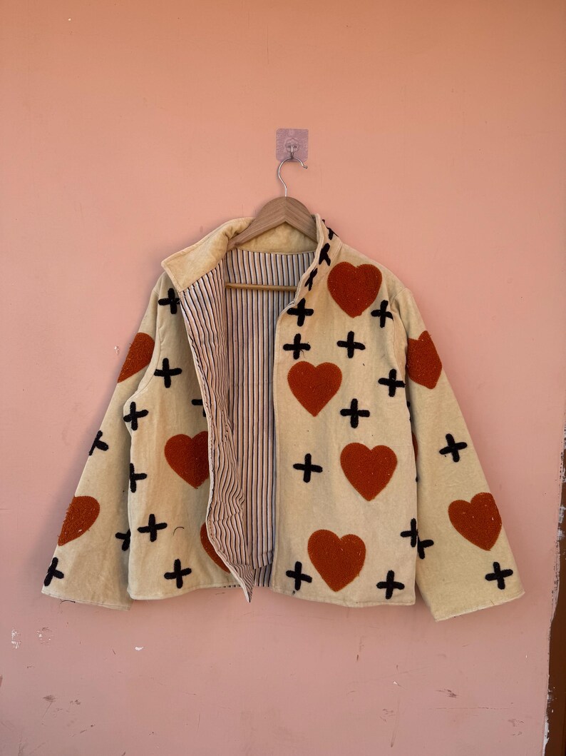 May include: Cream-colored jacket with red heart and black cross appliqu&eacute;s. The jacket has a collared neckline and a striped interior lining. It is hanging on a wooden hanger against a pink wall.
