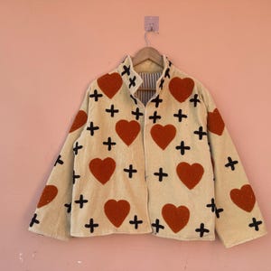 May include: Cream-colored jacket with a heart and cross pattern. The jacket features large red hearts and black crosses scattered across the fabric. The jacket is hanging on a wooden hanger.