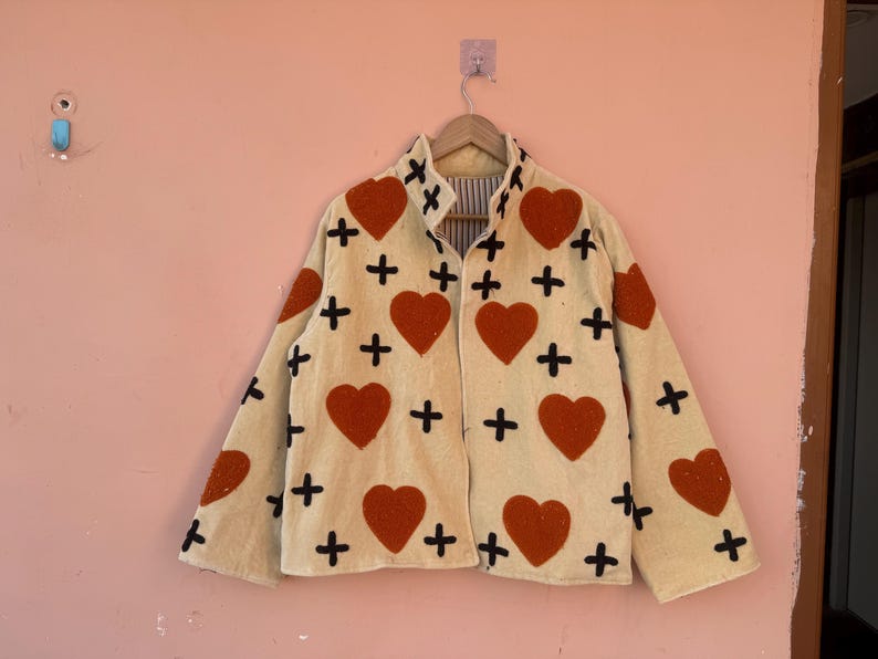 May include: Cream-colored jacket with a heart and cross pattern. The jacket features large red hearts and black crosses scattered across the fabric. The jacket is hanging on a wooden hanger.