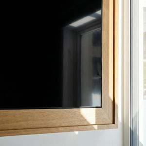 Könnte beinhalten: Ein Flachbildfernseher mit schwarzem Bildschirm ist in einen hellfarbigen Holzrahmen gefasst. Der Rahmen hat eine natürliche Holzmaserung und ist an einer weißen Wand montiert. Rechts ist ein Fenster zu sehen.