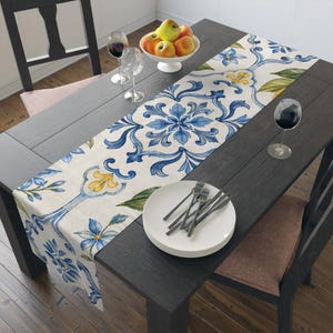 May include: A rectangular table runner featuring a repeating pattern of blue floral motifs, green leaves, and yellow accents on a cream-colored fabric. The runner is laid across a dark wooden table set with white plates, silverware, and wine glasses.