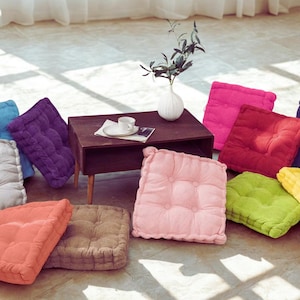May include: A selection of colourful square floor cushions in shades of pink, green, purple, and orange. The cushions are arranged on a light-coloured floor, with a small wooden table and a white vase in the background.
