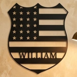 May include: Black metal shield-shaped wall art with a design resembling the American flag. The name "WILLIAM" is cut out at the bottom. The art piece is mounted on a beige wall. A desk with a book, notebook, and pen is in the foreground.