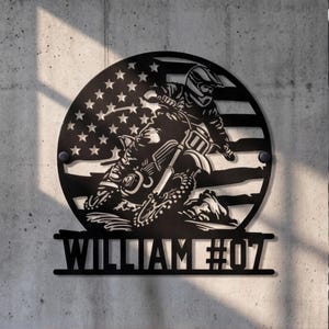 Puede incluir: Un letrero de pared circular de metal negro con un diseño recortado de un piloto de motocross en una moto de cross sobre un fondo de bandera estadounidense. Debajo del círculo, una barra horizontal muestra el nombre WILLIAM y el número #07.