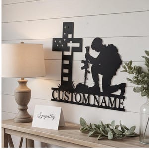May include: A black metal wall art piece featuring a silhouette of a soldier kneeling before a cross with stars and stripes cutouts. The base of the design includes the text CUSTOM NAME in bold capital letters, creating a memorial-style display.